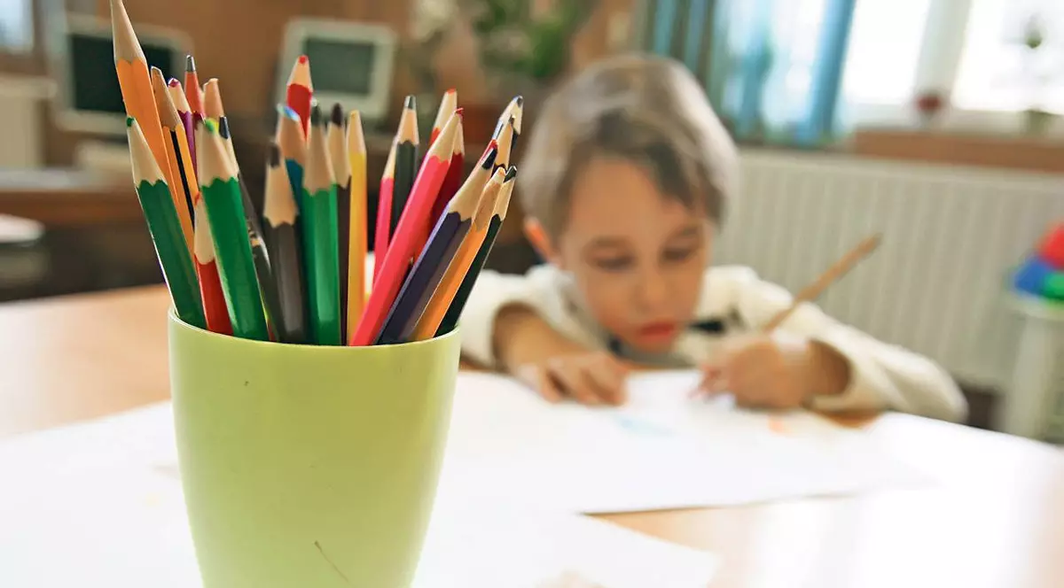 În prim-plan este un bol cu creioane colorate pus pe o masă, iar în fundal se vede un băiețel blond care colorează