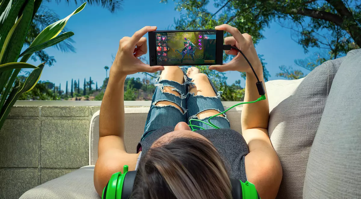 Producătorul de laptopuri și echipamente de gaming Razer a lansat Razer Phone, primul său smartphone, cu specificații de top, baterie mare și sunet THX. O fată stă pe spate pe o canapea și se joacă pe Razer Phone, având pe urechi o pereche de căști verzi Razer