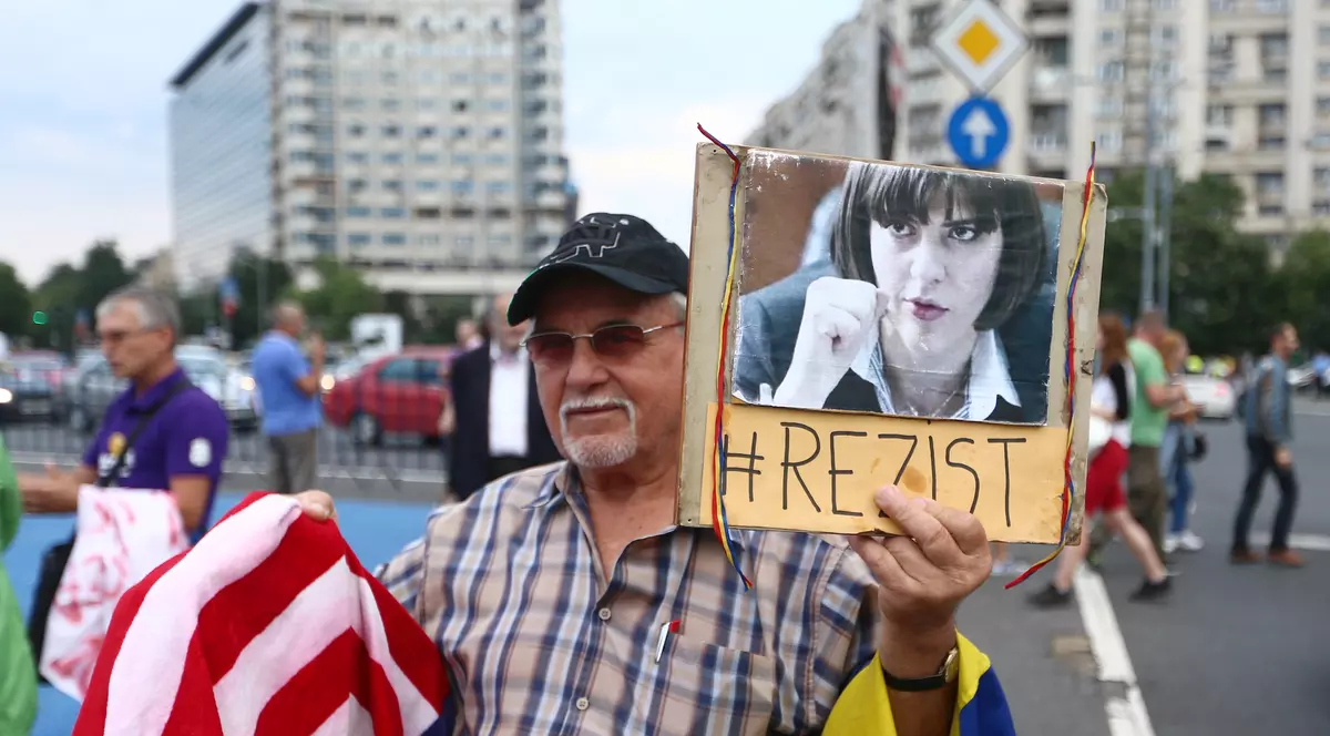 Protest în Piața Victoriei, pe 8 iulie. Un domn cu ochelri fumuri și cioc alb, șine pe brațul stâng drapelul României, iar în mâna stângă poza Laurei Codruța Kovesi, sub care scrie #Rezist, iar pe brațul drept drapelul SUA