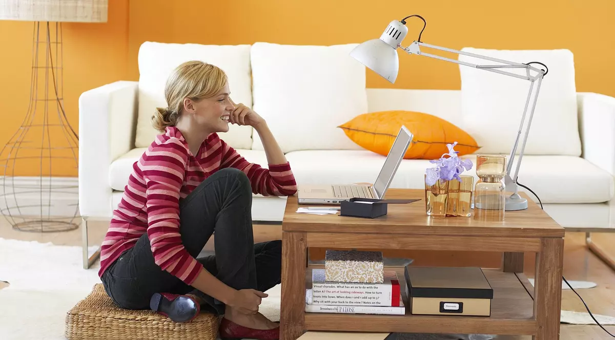 Eurostat: Românii și bulgarii, pe ultimele locuri din UE la munca de acasă. Femeie pe parchet, în fața unui laptop, cu o canapea și o lampă pe fundal