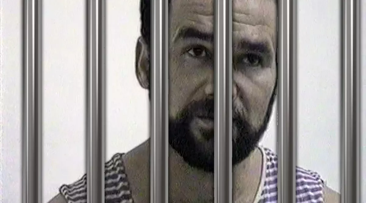 Un bărbat cu barbă, îmbrăcat în maiou cu dungi, este în spatele gratiilor