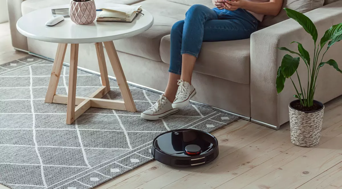 Reduceri la produsele de casă în oferta AloShop de Black Friday 2022, azi, 8 noiembrie - Imagine cu o tânără aşezată pe canapea, privind un telefon. În imagine se mai observă un robot de aspirare, un covor şi o masă cu o floare pe ea