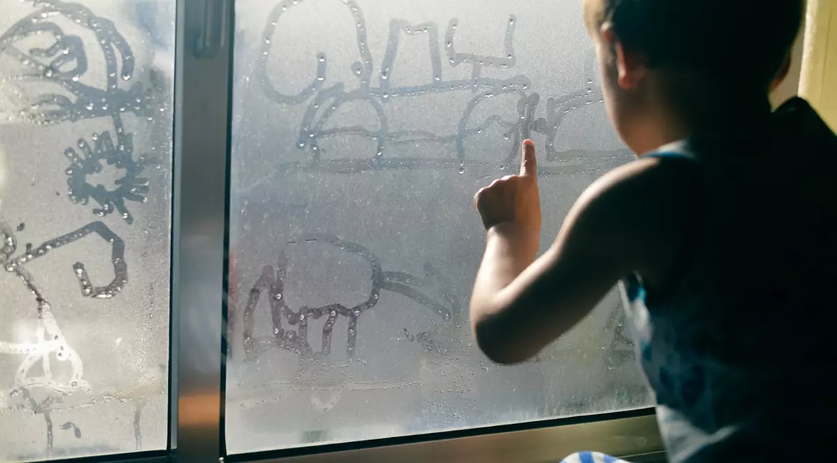 Ce este condensul, de ce apare și cum îl putem evita - Imagine cu un copil care desenează cu mâna pe un geam aburit