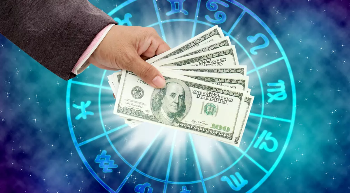Zodii cu noroc la bani în această toamnă - Imagine cu o mână care ţine mai multe bancnote de câte 100 de dolari, deasupra uni planşe cu semnele zodiacale