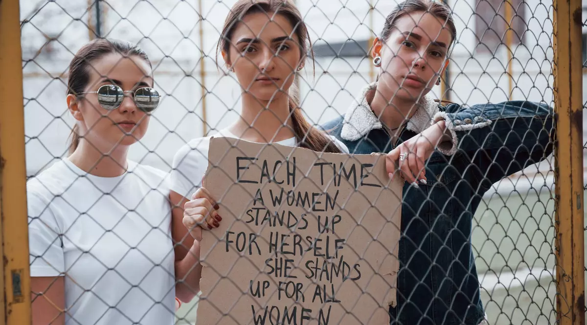grup de femei care au un mesaj feminist scris pe un carton, in spatele unui gard