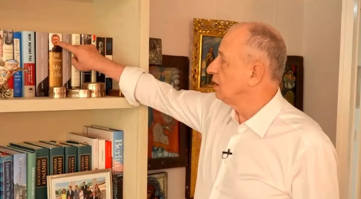 Mircea Geoană, îmbrăcat cu o cămașă albă, în timp ce arată cu degetul către o carte din biblioteca sa