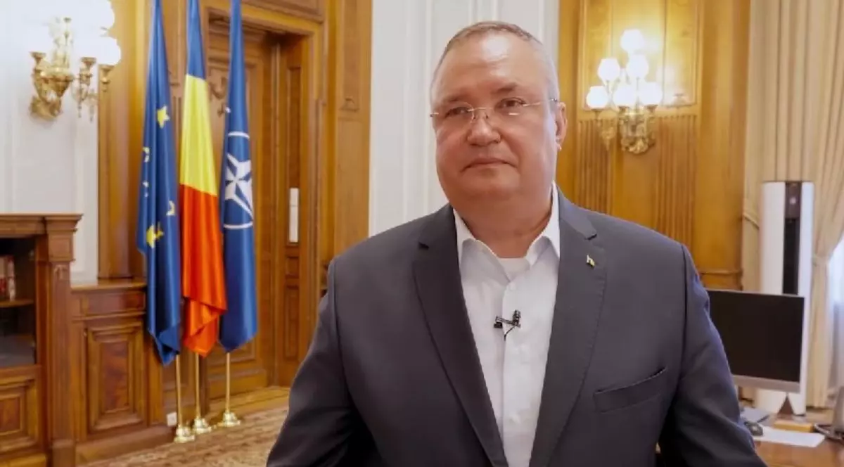 Nicolae Ciucă, îmbrăcat în costum, într-un birou de la Parlamentul României, pe fundal cu pereți acoperiți cu lemn și steagurile Uniunii Europene, al României și cel al NATO