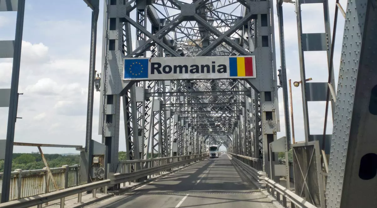Podul Giurgiu-Ruse, podul Prieteniei, văzut dinspre Giurgiu. Pe șosea se află un TIR alb, iar deasupra ramei metalice ale podului se observă un semn de circulație cu textul „România”, steagul Uniunii Europene și steagul României