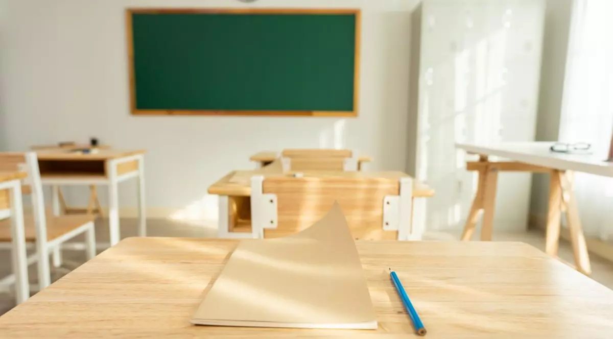 O sală de clasă cu tablă și bănci goale, în prim-plan un caiet și un creion albastru pe masă. Foto: Shutterstock