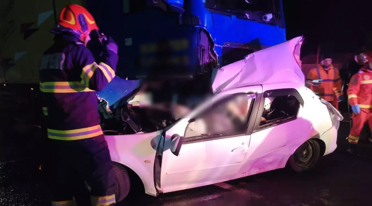 Mașina de culoare albă, lovită în lateral de camionul de culoare albastră, în timp ce pompierii încearcă să scoată victimele din interior