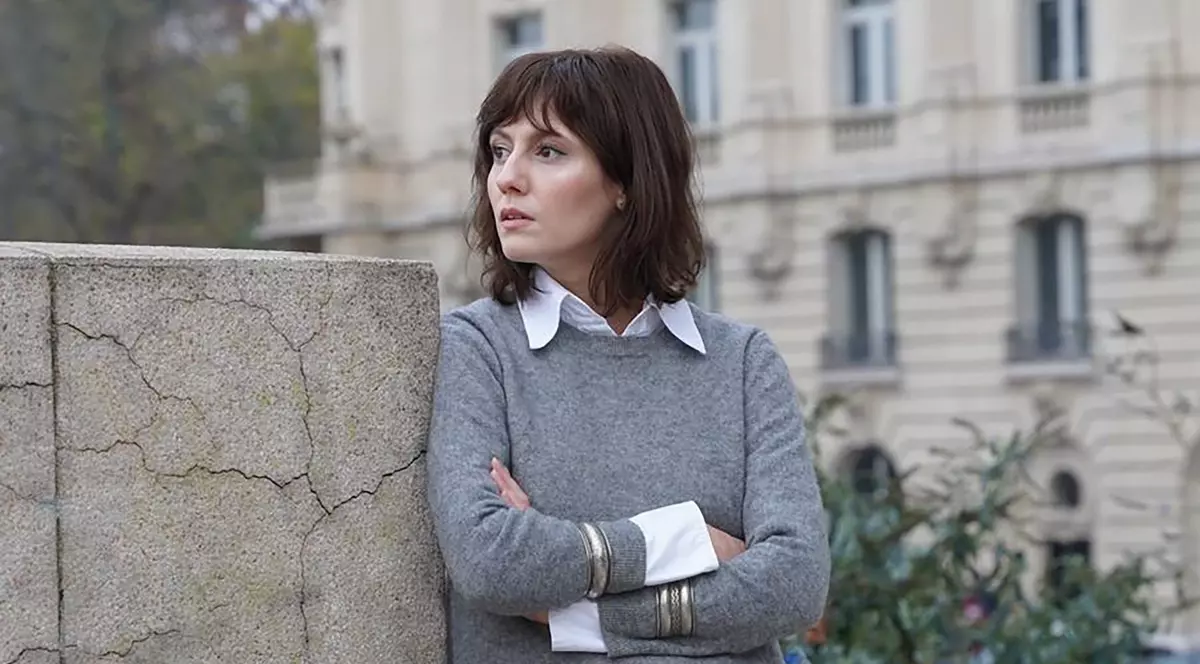 Ada Galeș, îmbrăcată cu o bluză de culoare gri care acoperă o cămașă de culoare albă, sprijinită de un perete