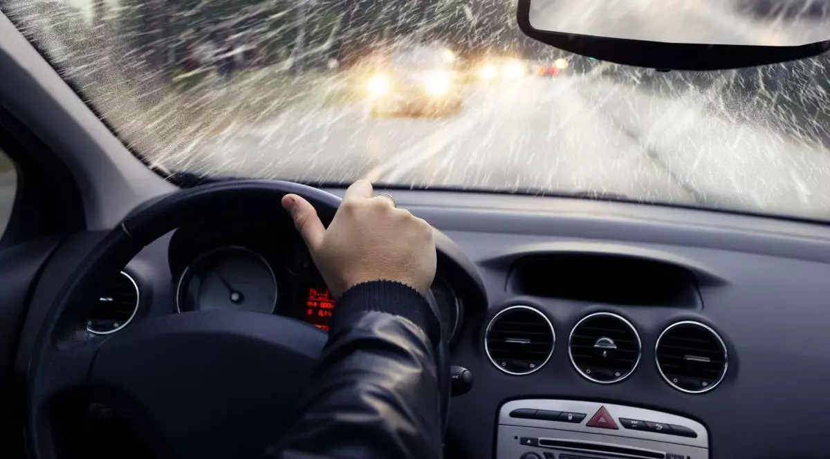 Un bărbat îmbrăcat cu geacă este cu mâna pe volanul mașinii și conduce într-o zi de iarnă în care afară se vede lapoviță