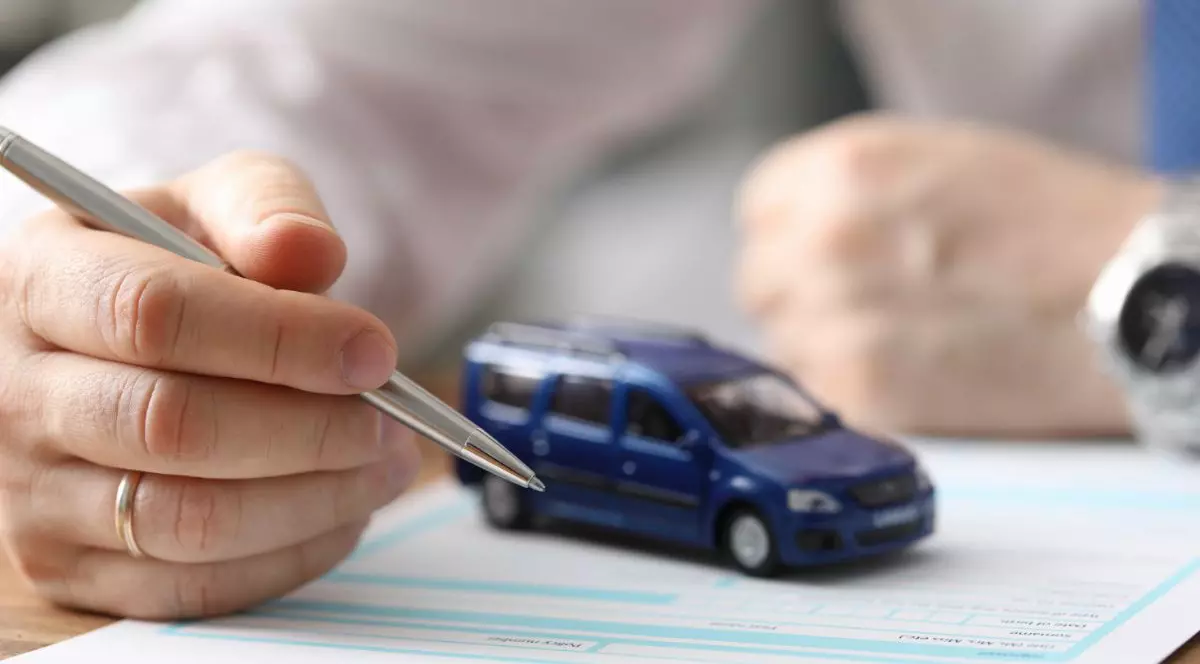 barbat cu un pix în mână care semnează o asigurare RCA. O macheta cu o mașină mică albastră este așezată pe foile din fața bărbatului