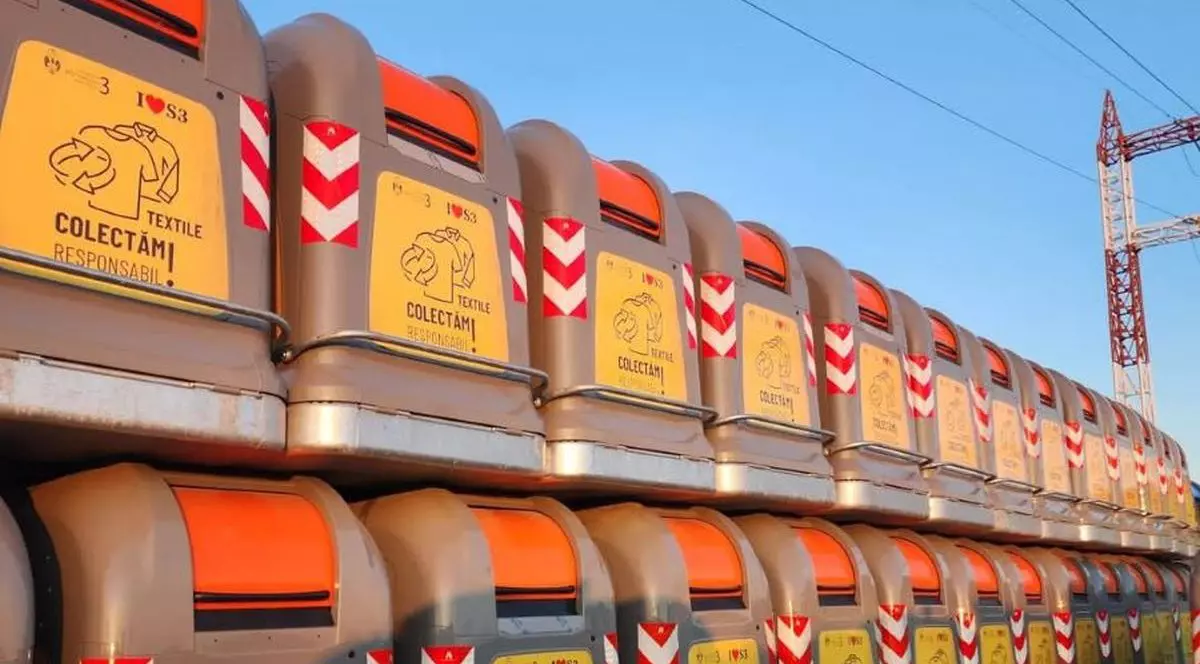 Mai multe containere gri pentru colectarea hainelor vechi, puse unele peste altele, se văd pe o stradă