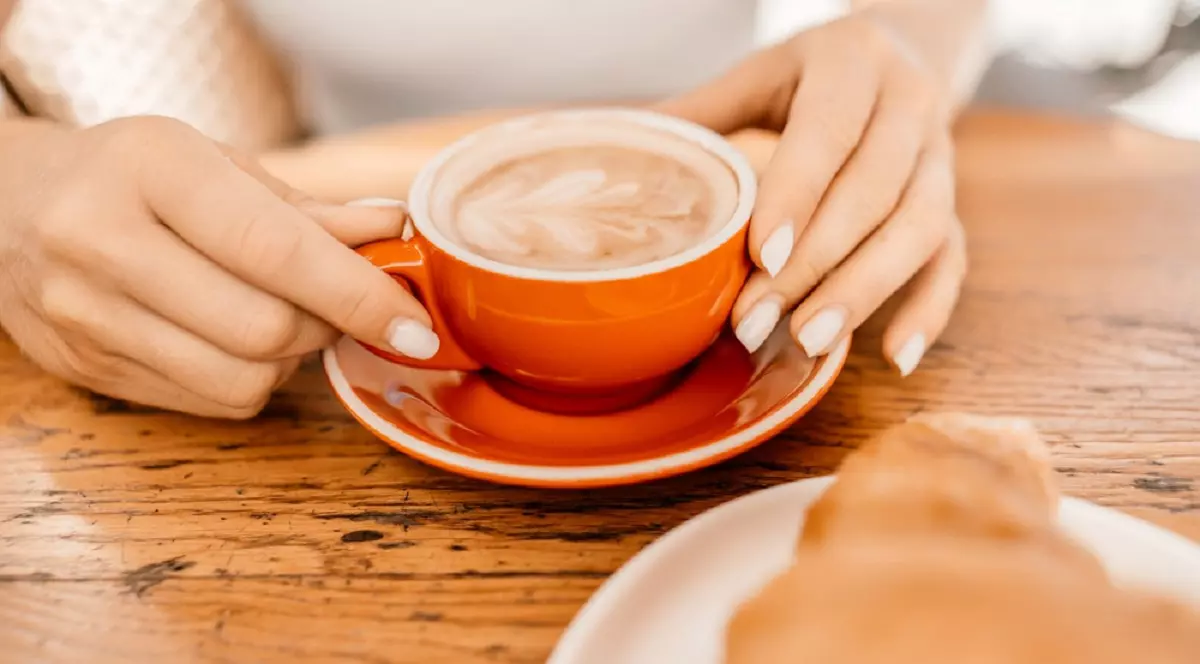 O femeie sta la o masa de lemn si își tine mainile pe o ceasca rosie plina cu cafea cu lapte