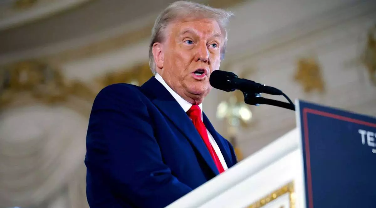 Donald Trump, îmbrăcat într-o cămașă albă cu cravată roșie și sacou albastru, vorbește la microfon, de la tribună