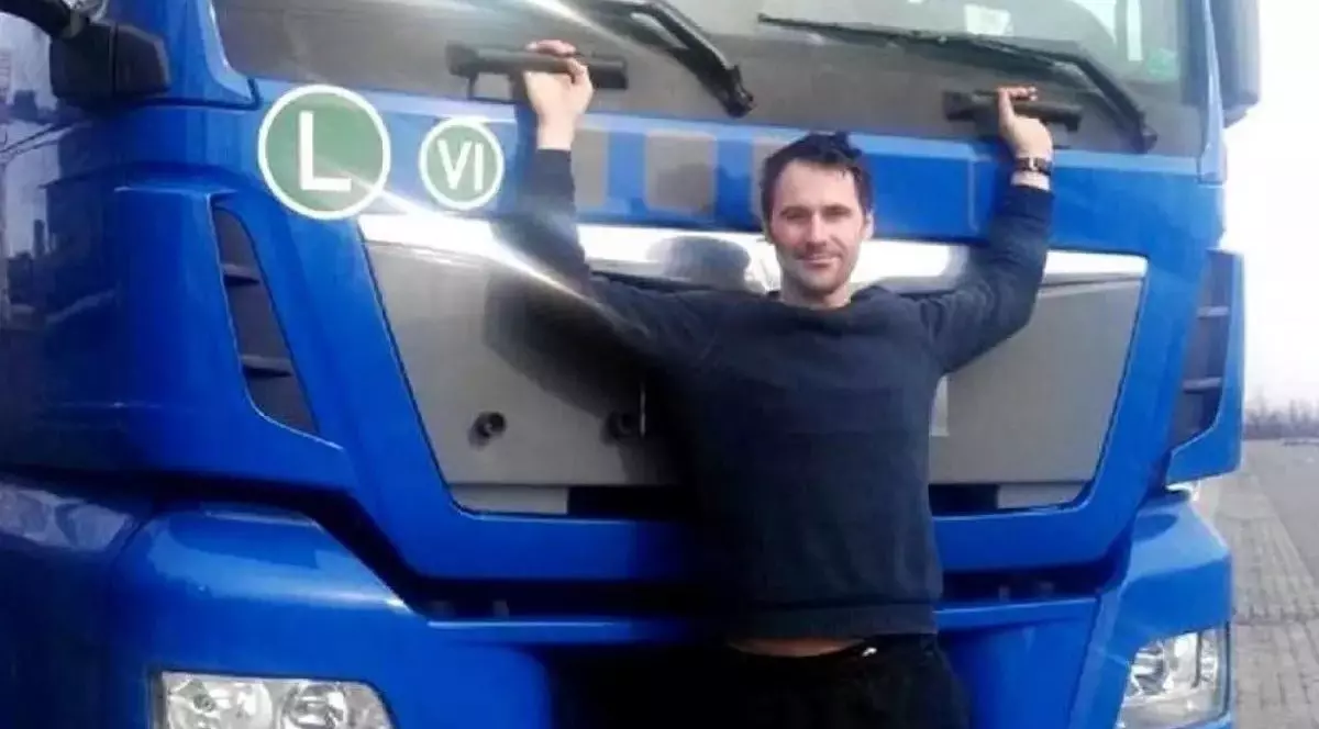 Un bărbat se ține cu mâinile de cabina unui camion albastru, stând în picioare în fața ei
