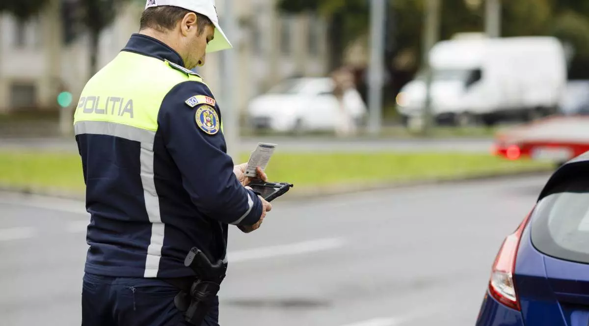 politist imbracat in uniforma in timpul unui control de rutina al unui sofer oprit in trafic, langa este o masina albastra oprita