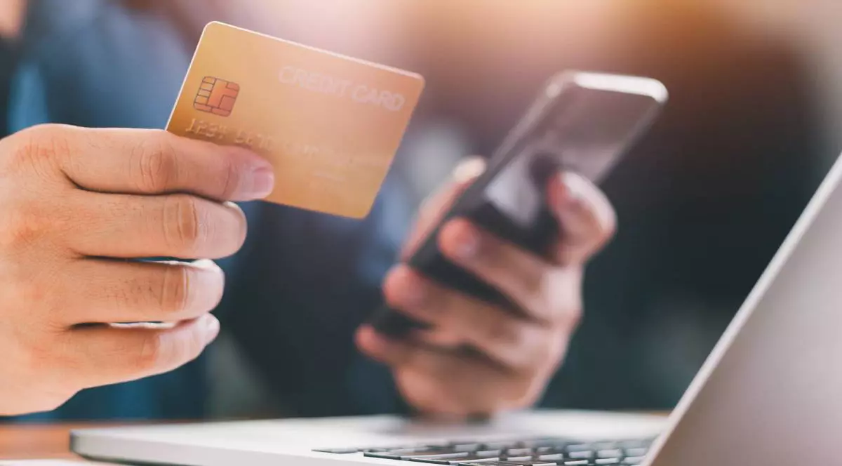 barbat care tine intr-o mana un card bancar si in cealalta un telefon mobil, in fata are un laptop