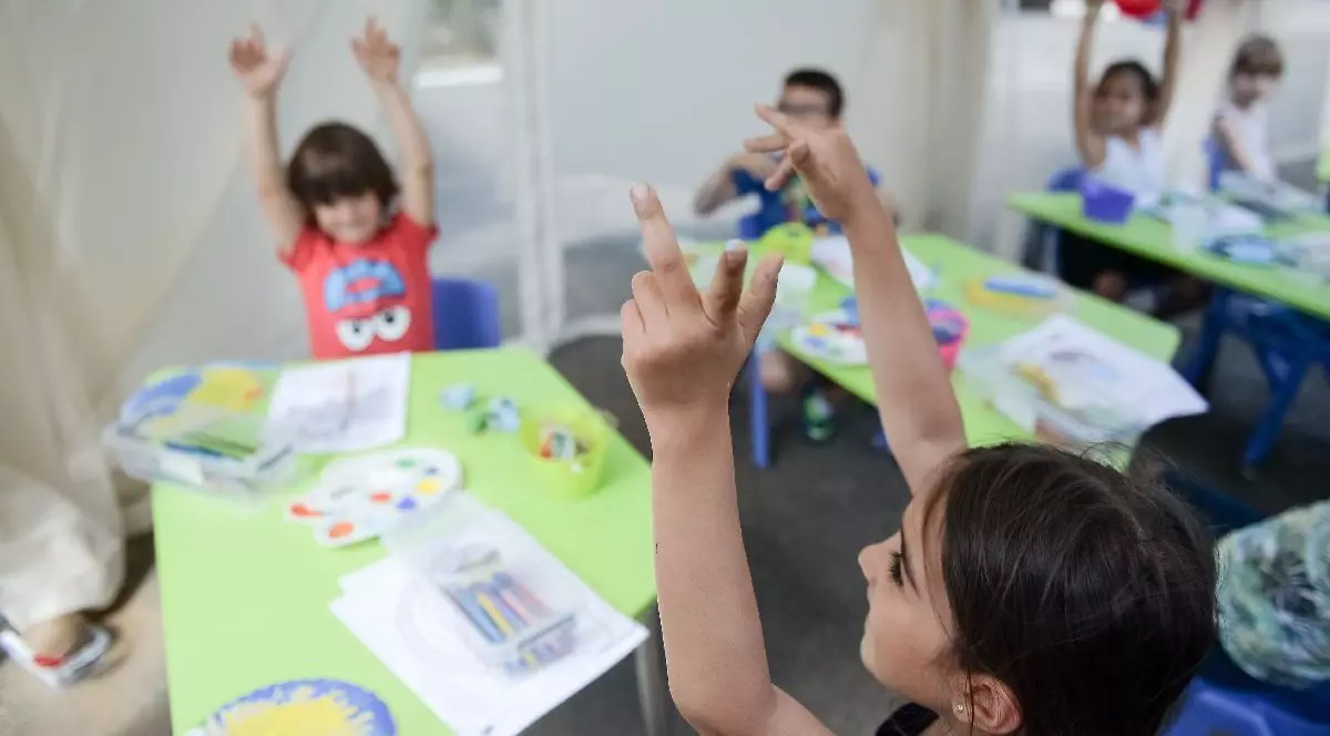 copii așezați la măsuțe verzi, într-o sală de clasă.