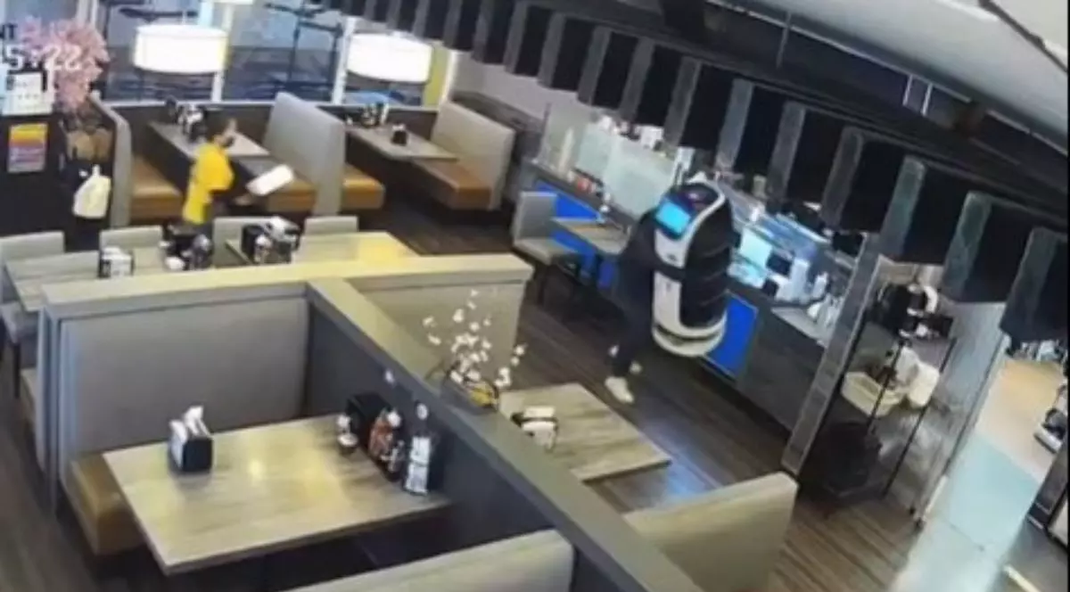 un bărbat cu șapcă, care îi acoperă fața, care cara un chelner robot, în timp ce o angajată îmbracată într-o cămașă galbenă fuge după el