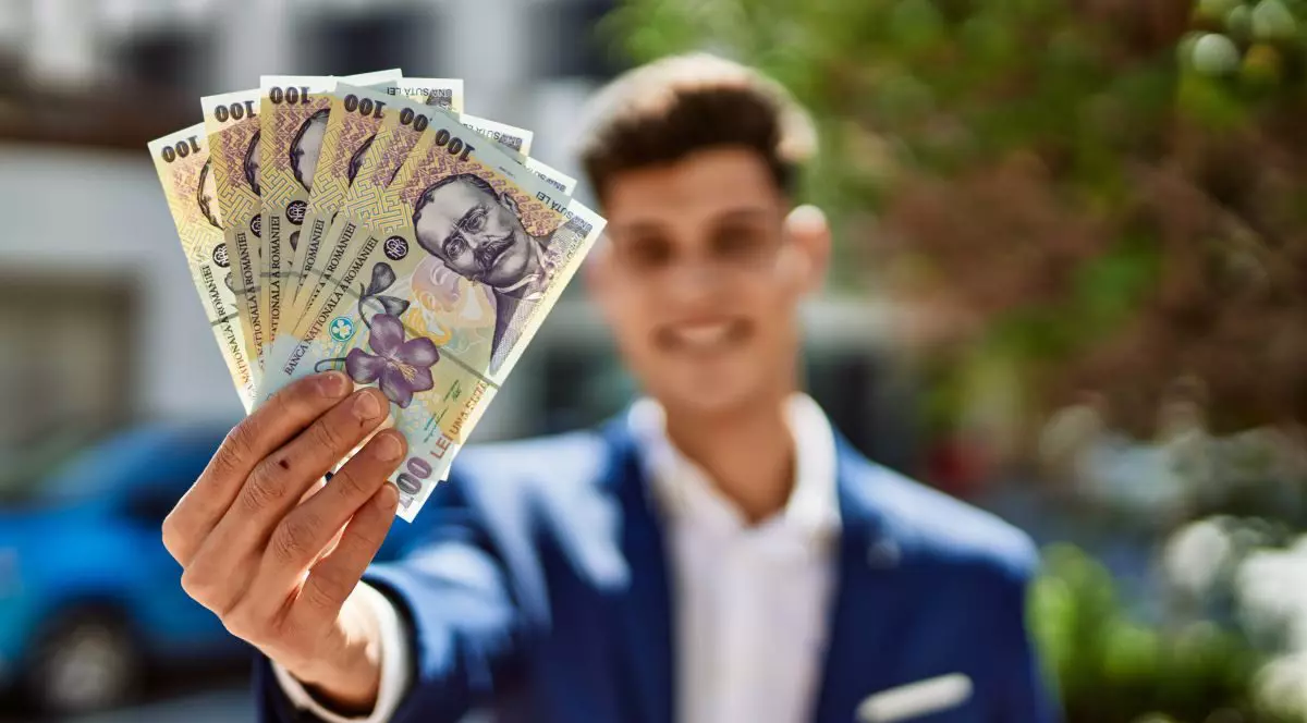 Un bărbat îmbrăcat la costum și cu fața blurată ține în mână mai multe bancnote de 100 de lei și le arată