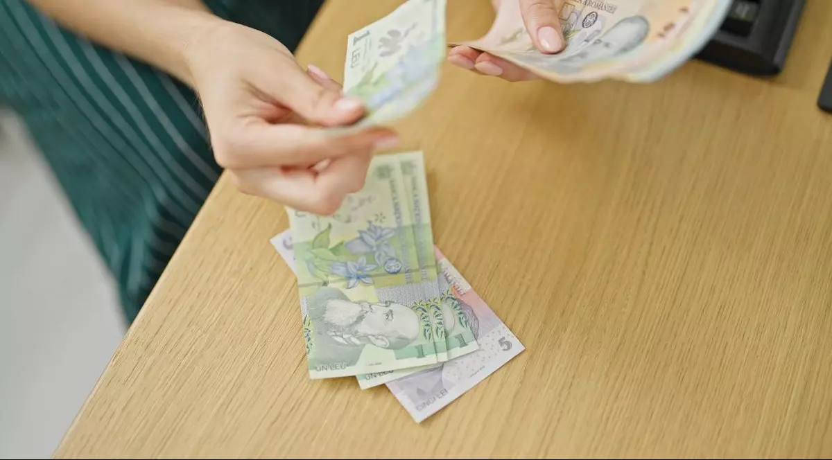 O femeie are în mână niște bani românești pe care îi numără și pune bancnotele pe biroul de lângă ea, unde se vede și o tastatură. Foto: Shutterstock