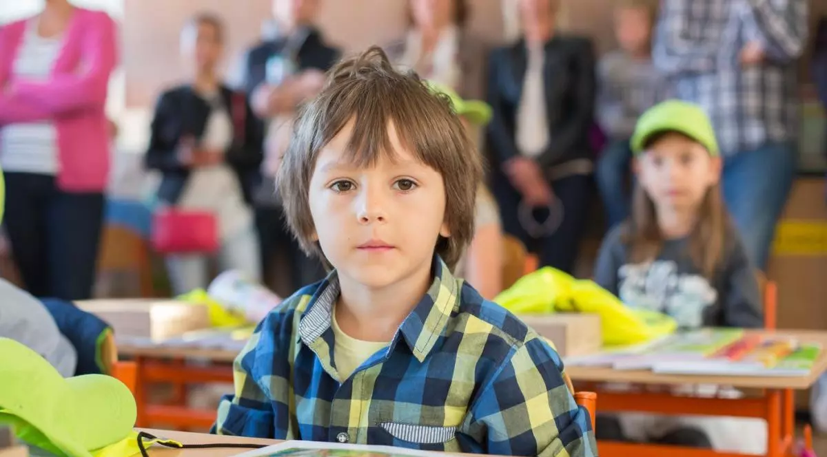 Un băiat la clasa pregătitoare, îmbrăcat cu o cămașă pepită, stă în bancă în prima zi de școală. În jurul lui sunt alți copii, iar în spate se văd părinții acestora