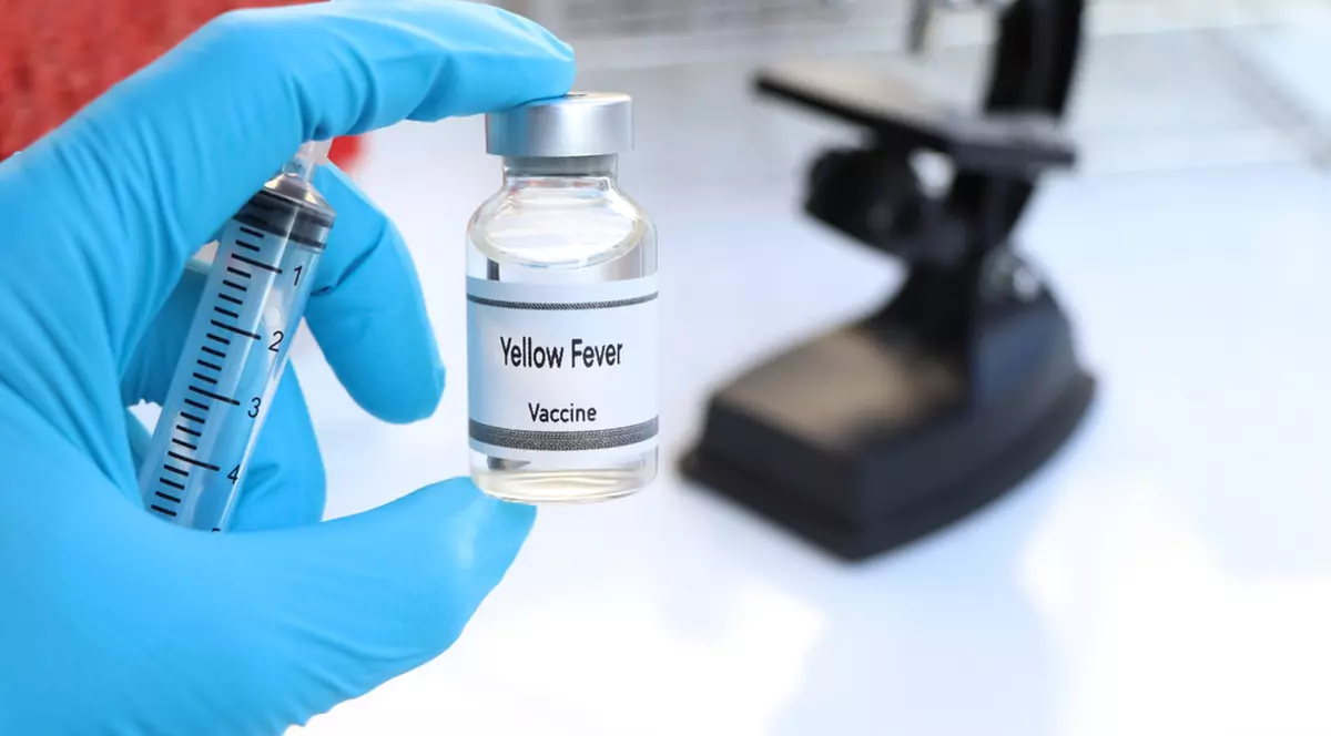 Ce este febra galbenă. Cum se transmite, simptome, tratament şi prevenţie - Imagine cu o persoană care ţine în mână un flacon de vaccin pentru febra galbena, având în fundal un microscop şi o seringă.