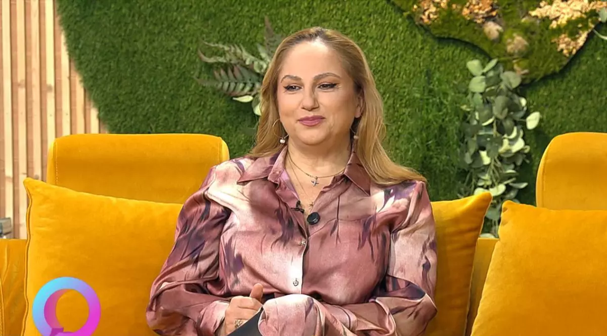 Astrologul Cristina Demetrescu, îmbrăcată cu o bluză maro, stă pe o canapea portocalie, cu un decor verde în spate