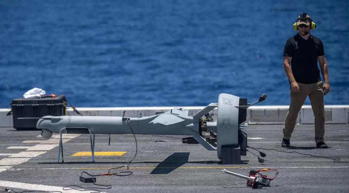 dronă de tip V-BAT aflată pe o platformă de aterizare pe un vas militar. Lângă dronă, un operator echipat cu căști de protecție și ochelari de soare supraveghează pregătirile. Pe platformă sunt vizibile mai multe echipamente, inclusiv o cutie de transport și diverse cabluri,