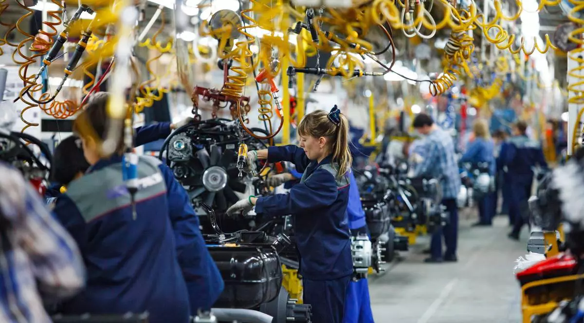 fabrica auto unde mai mulți muncitori lucrează la montajul componentelor vehiculelor. În prim-plan, o femeie îmbrăcată în echipament de protecție