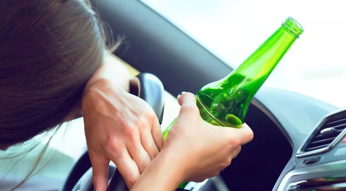 Femeie băută la volan. Stă cu capul pe volan, iar în mâna dreaptă are o sticlă cu alcool