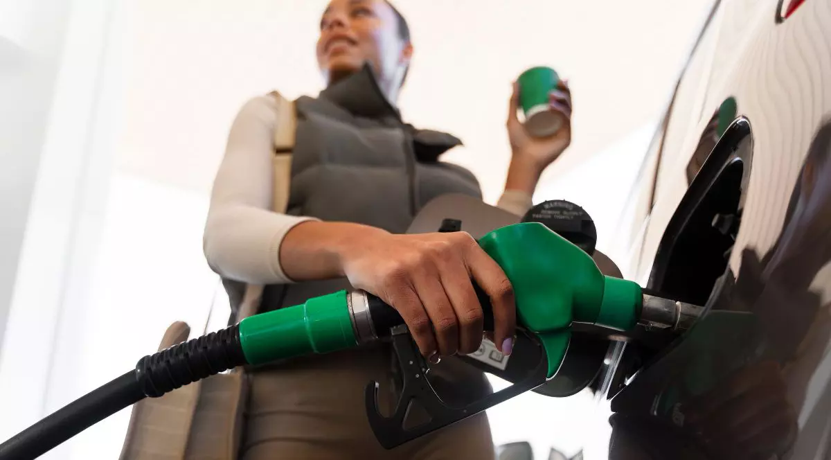 femeie care își încarcă automobilul cu combustibil, în timp ce ține un pahar cu cafea în mână