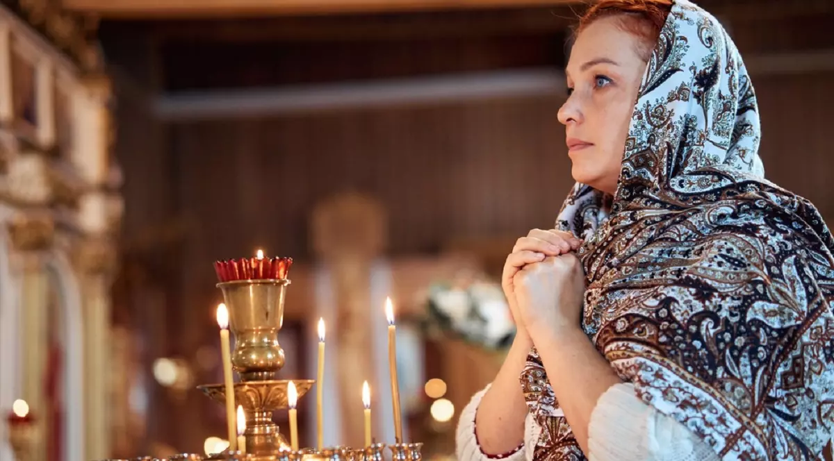 Ce simbolizează rugăciunea „Tatăl Nostru” - Imagine cu o femeie care se roagă într-o biserică, lângă un suport cu lumânări aprinse