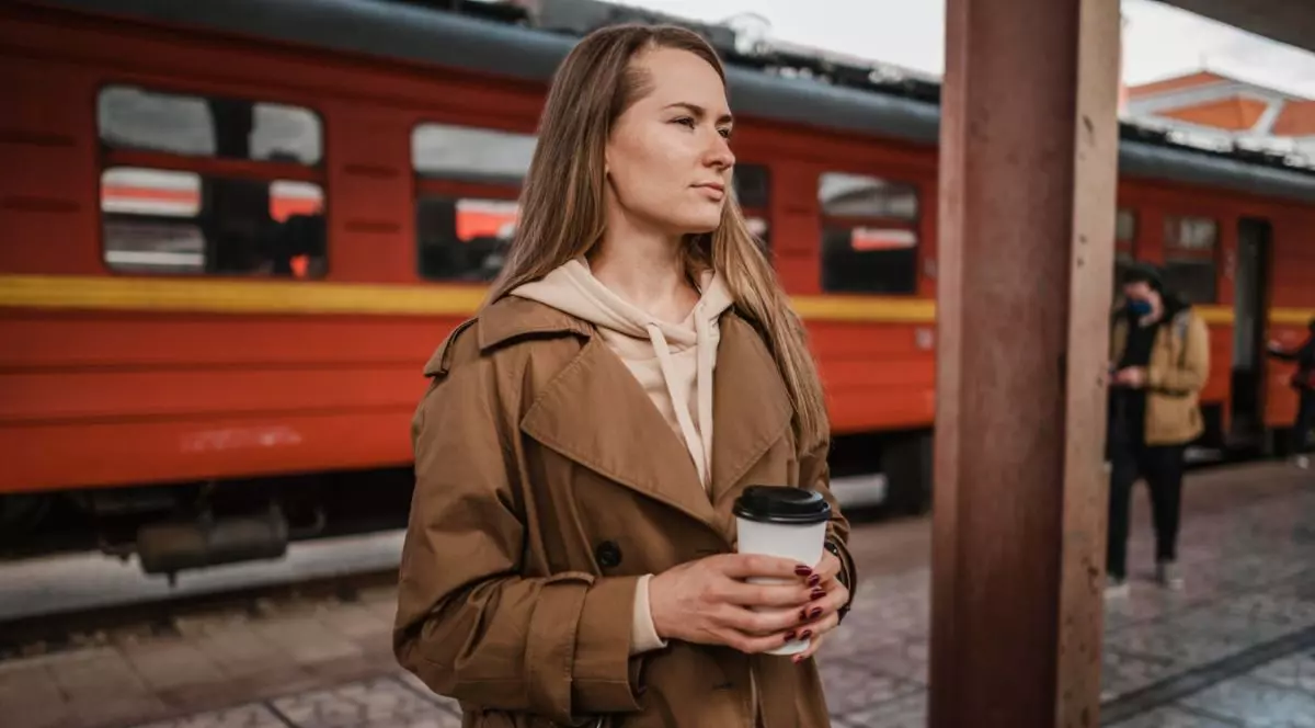 femeie care ține un pahar de cafea într-o gară, cu un tren roșu pe fundal și alți călători