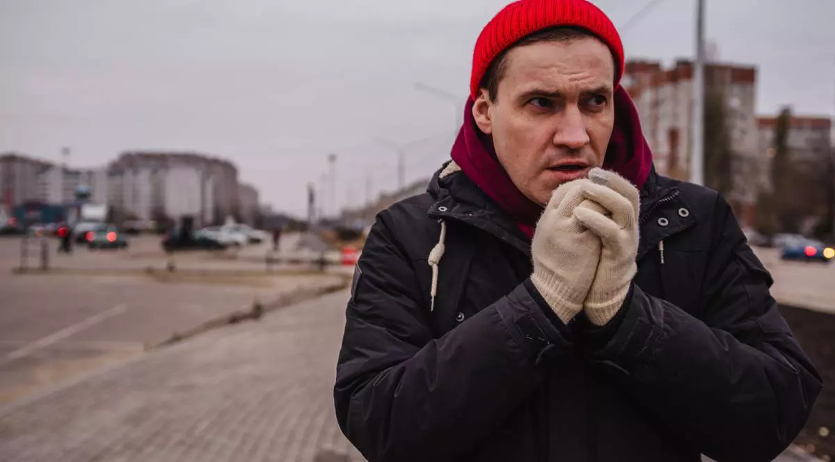 barbat imbracat cu geaca si fes pe cap, manusi. in timp ce isi sufla in pumni de frig