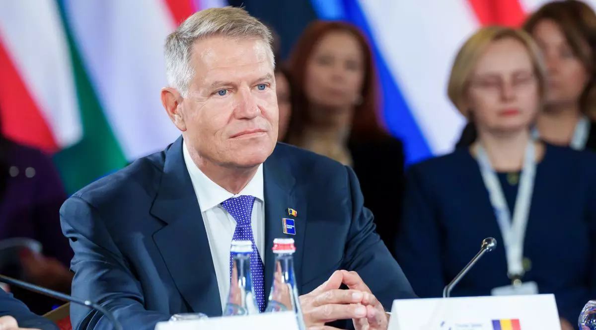 Președintele Klaus Iohannis, îmbrăcat cu costum și cravată cu picățele, stă gânditor la o masă, unde este un cartonaș cu steagul României, iar în jurul lui se văd ușor neclar alte persoane