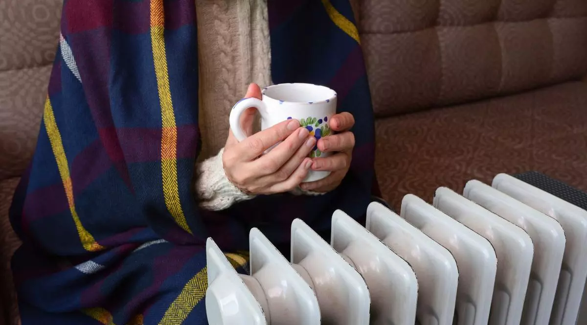 O femeie cu pulover cu guler și un șal pe ea stă înfrigurată pe canapea, cu o cană de ceai în mână, lângă un calorifer