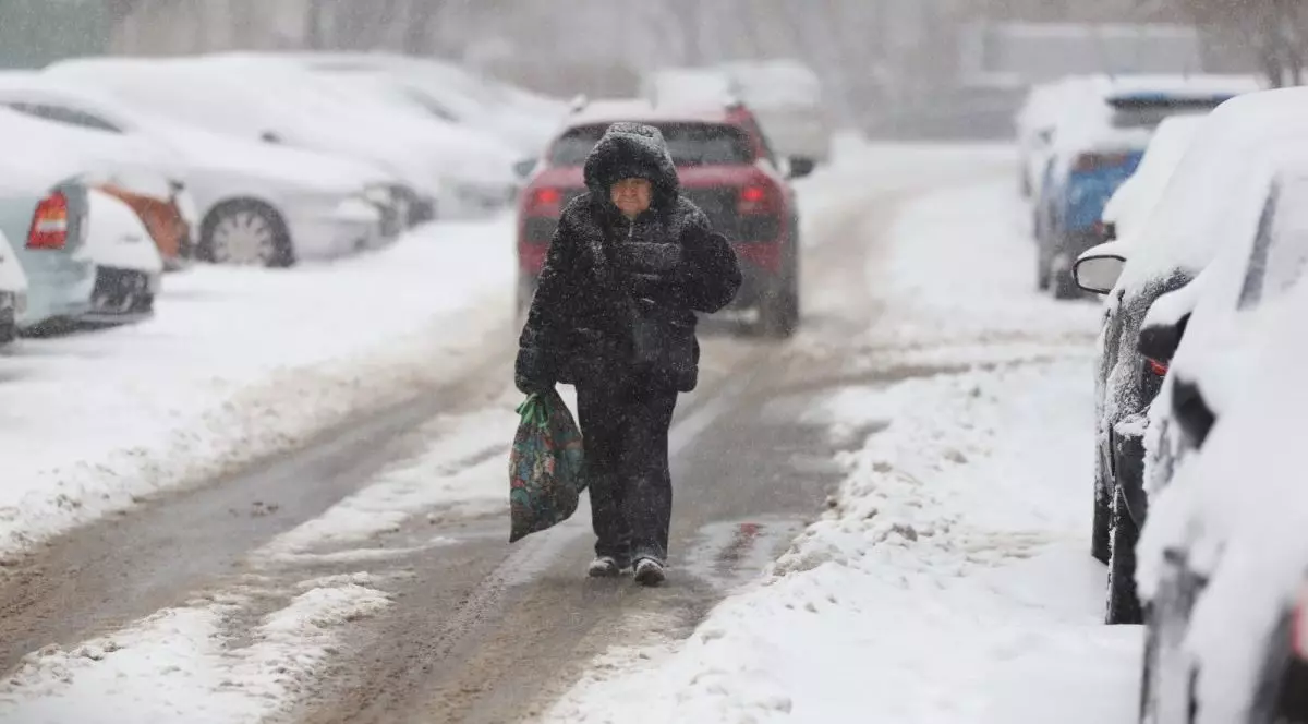 O femeie bătrână îmbrăcată în negru, cu glugă pe cap și o sacoșă în mână, merge pe stradă, pe o porțiune care este deszăpezită, pe unde trec mașinile