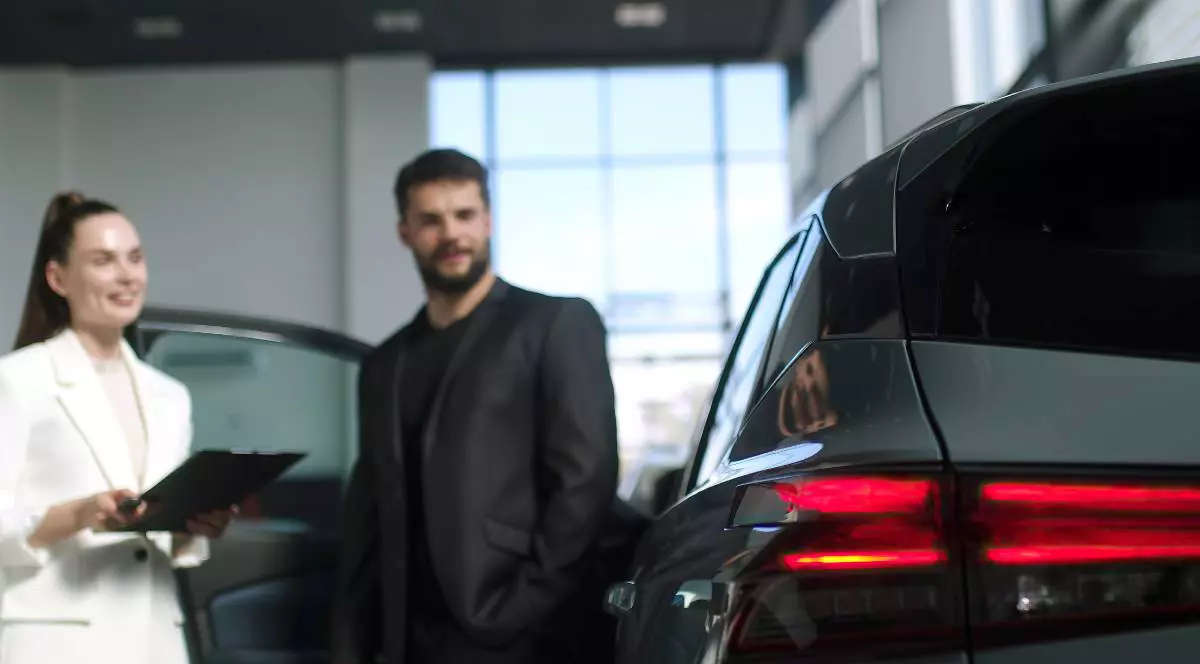 un barbat imbracat la costum, posibil client, alaturi de o femeie care tine in mana cateva foi, posibil informatii despre masina de langa cei doi