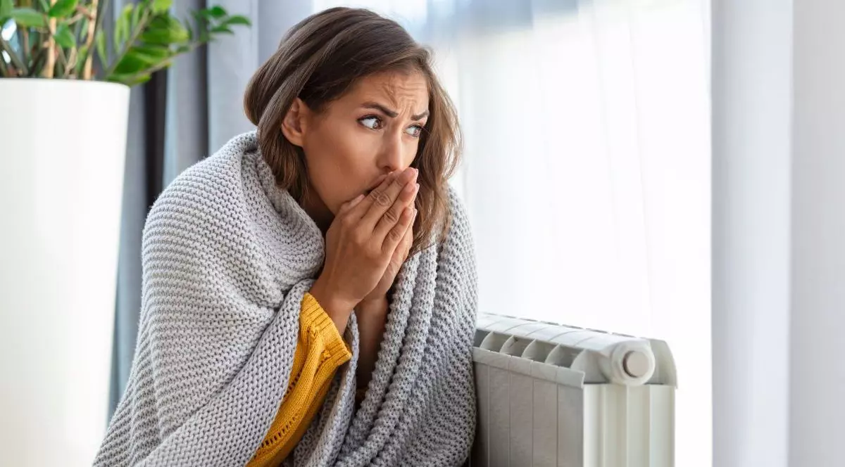 O femeie cu multe haine pe ea stă acasă lângă un calorifer alb, se uită pe fereastră și își suflă în mâini de frig