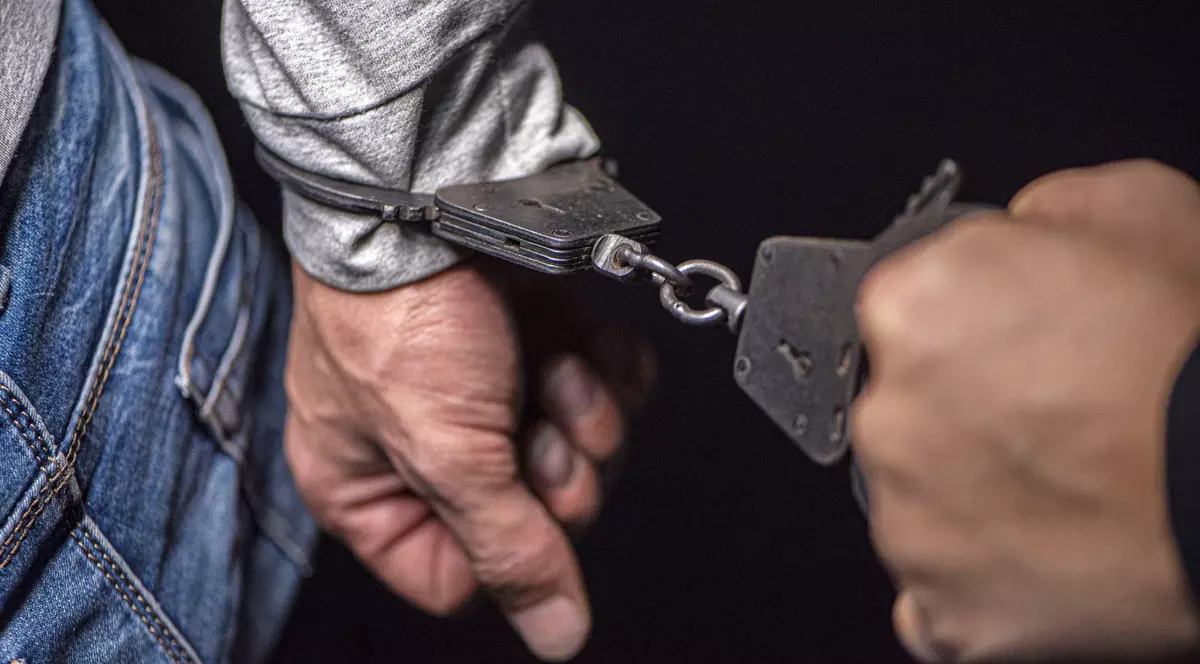 close-up al un bărbat cu mâinile în cătușe, îmbrăcat într-un hanorac gri și jeanși albaștri