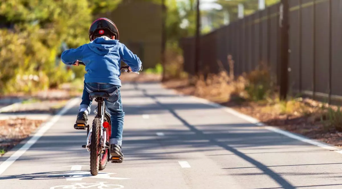 Un copil pe bicicletă, fotografiat cu spatele, merge pe trotuar într-o zi cu soare