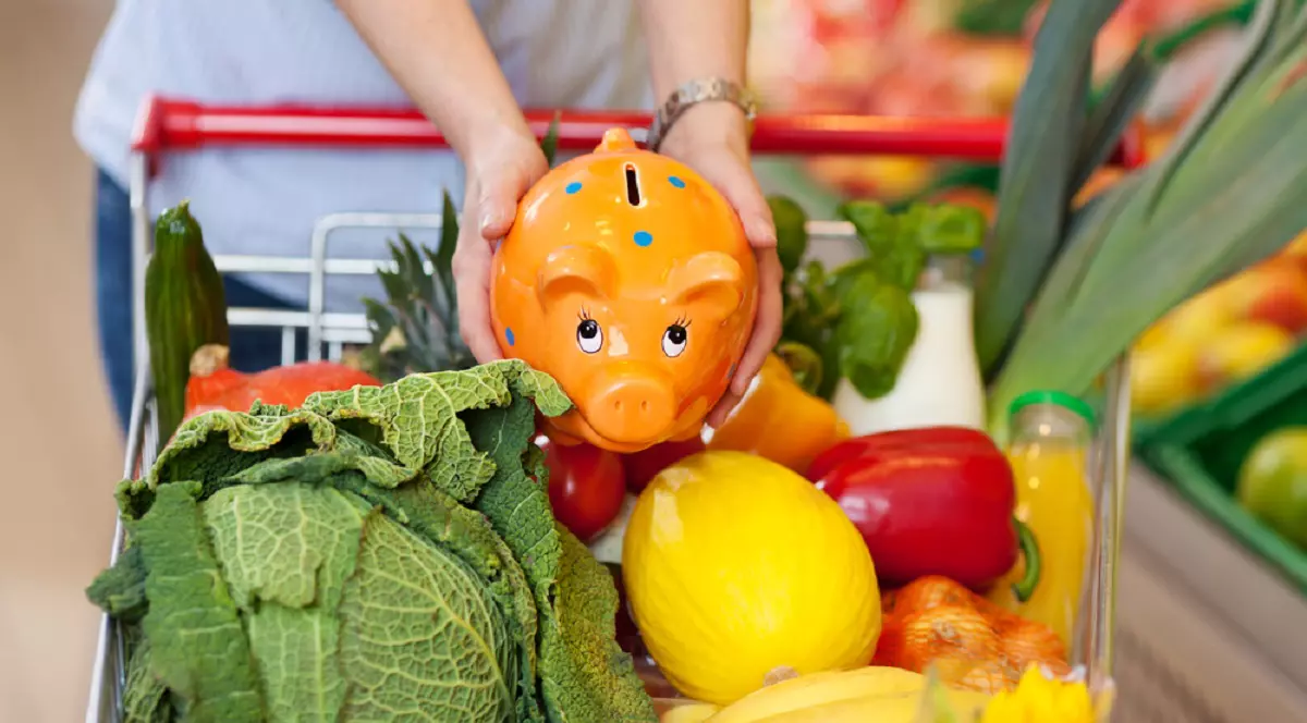 Ce este metoda „6 la 1” prin care poți să faci economie la cumpărături - Imagine cu o femeie care ţine cu ambele mâini o puşculiţă "porcuşor" ceramică, desupra unui cărucior de cumpărături la supermarket