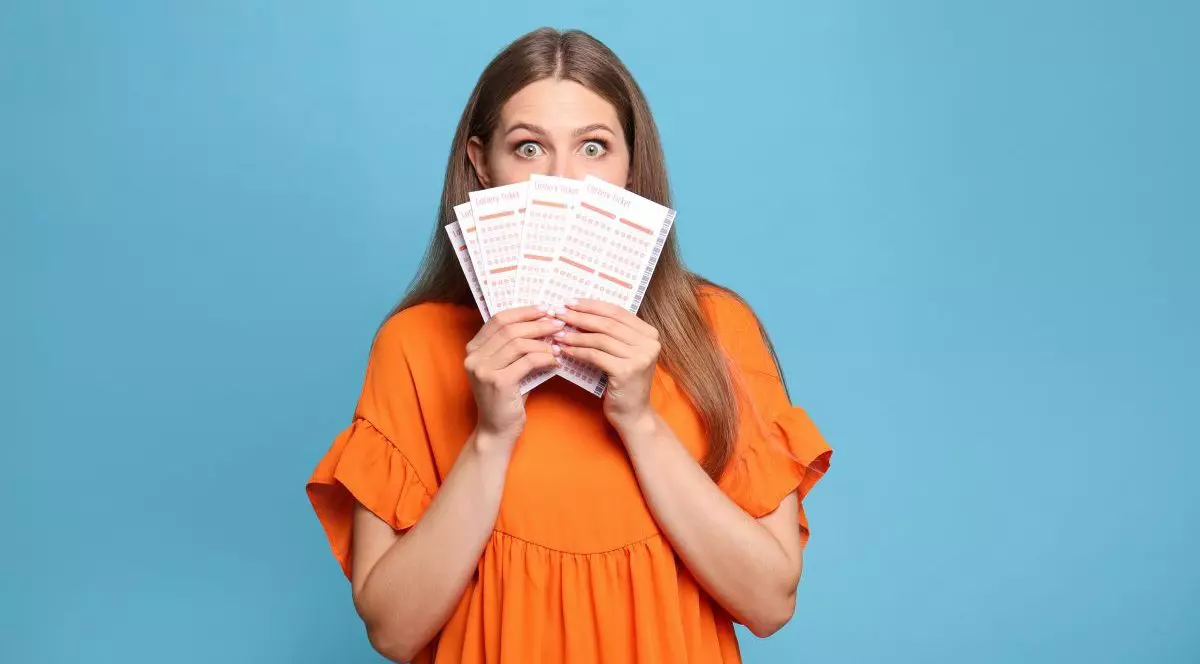 O femeie îmbrăcată în rochie portocalie, pe un fundal bleu, ține în mâini câteva bilete la jocurile de noroc și își acoperă o parte din față cu ele, fiind foarte surprinsă