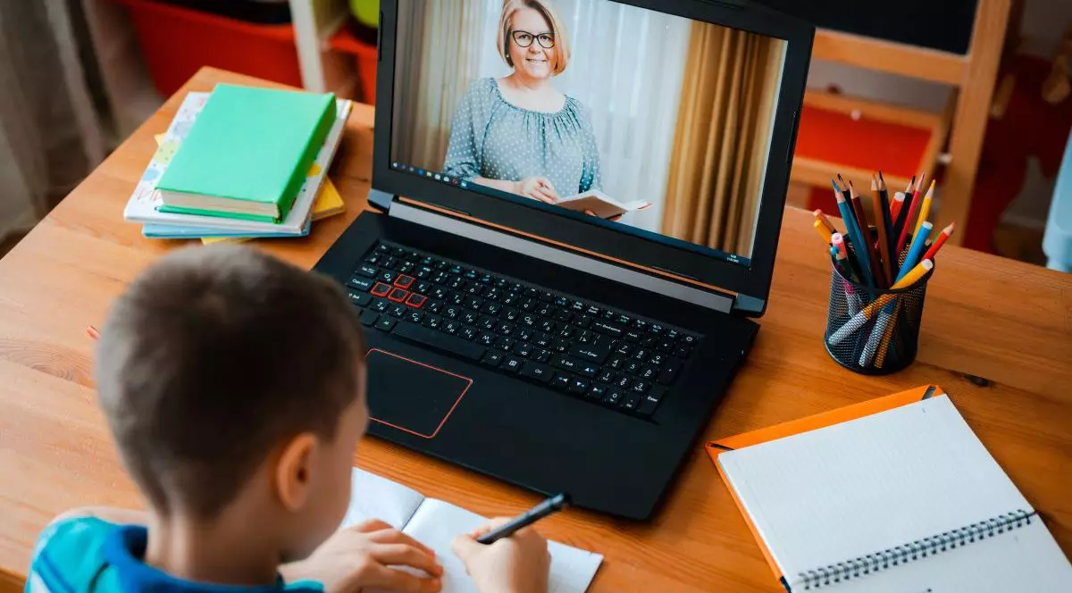 Un elev care stă cu spatele scrie pe caiet, iar în fața lui este un laptop deschis pe care comunică cu profesoara, care se vede pe ecran