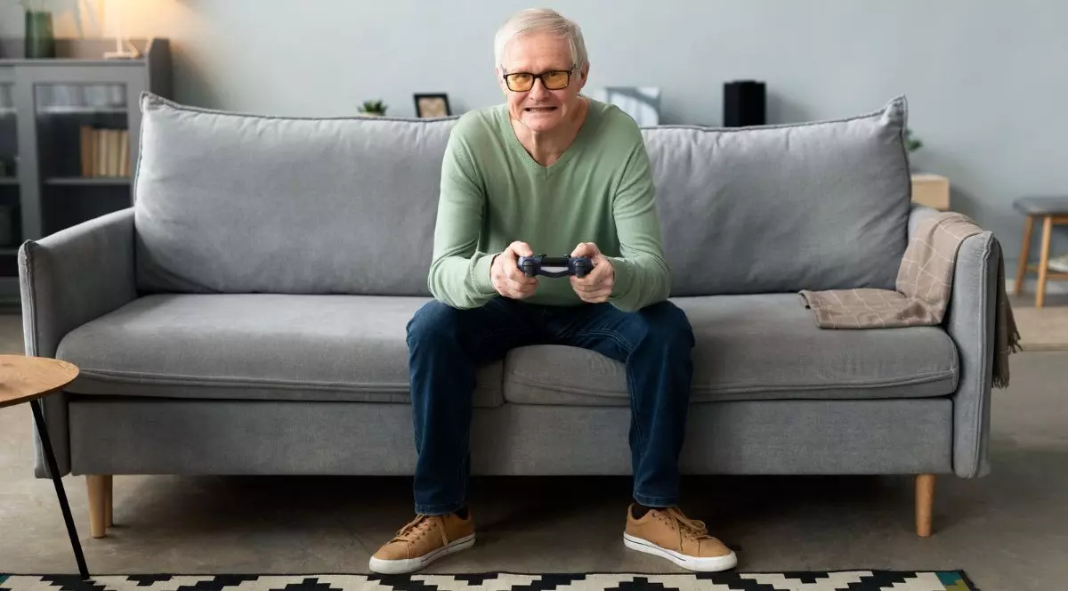 bărbat în vârsta așezat pe o canapea gri, care se joacă jocuri video cu ajutorul unui controller