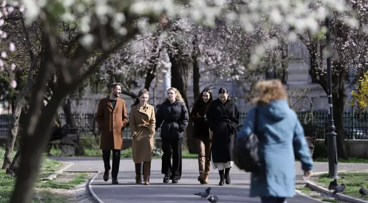 Cinci tineri îmbrăcați cu geci și paltoane merg pe o alee din parcul Cișmigiu din București, în fațalor sunt doi porumbei, iar în jur se văd copaci înfloriți