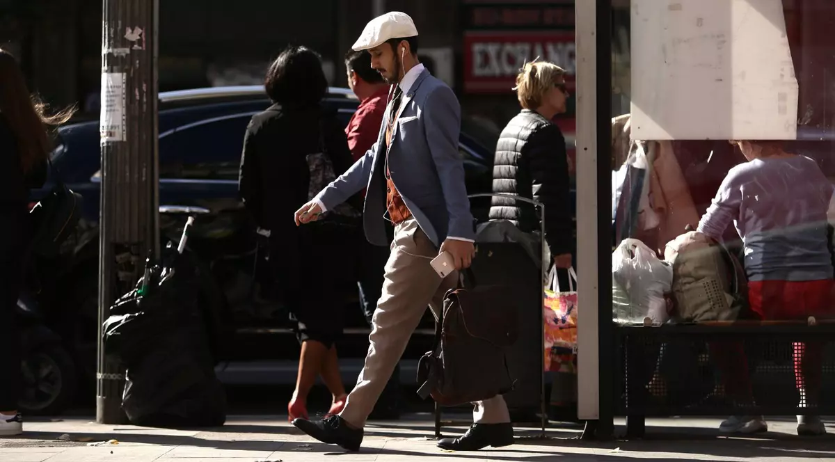 Mai mulți oameni merg pe stradă într-o zi însorită, iar în centru se vede un bărbat tânăr îmbrăcat la costum, cu o șapcă albă, căști și un telefon în mână. Foto: Libertatea/Dumitru Angelescu